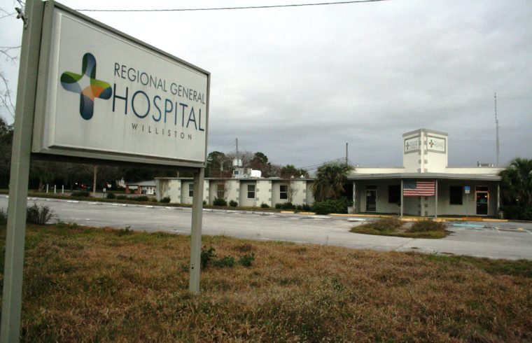 The front yard sign of Regional General Hospital stands in an overgrown yard. The hospital itself is beginning to look abandoned.