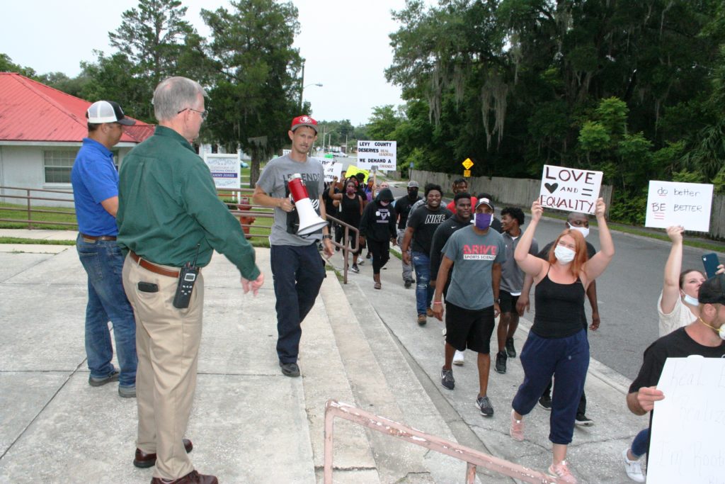 Sheriff Bobby McCallum waves to protesters as they pass by the front of the Levy County Courthouse steps. Standing at his side is County Commission Chairman Matt Brooks.