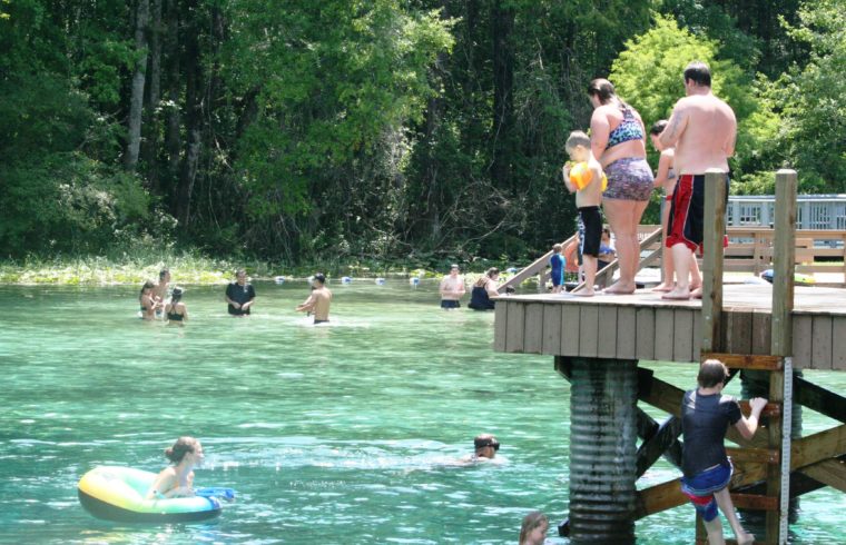 Two adults and small children stand on the diving dock at Blue Springs Park taking in the beautiful scene below and around them. Some of their children were more adventurous than others and jumped in the water feet first with no hesitation. Others had to study the situation first. Visitors can be seen playing water ball in the distance.