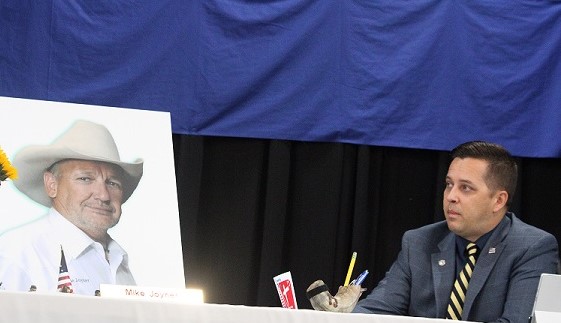 County Commissioner Matt Brooks is seated next to a poster of his friend, the late County Commissioner Mike Joyner, as he holds a decal suggested by Joyner just weeks before his passing. The decal said, "Stand for the flag, kneel for Christ."