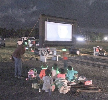 A small group of children cuddle up on the basketball court to watch the movie. They are wearing luminous, wireless headphones to listen to the movie as they watch on an inflatable screen.